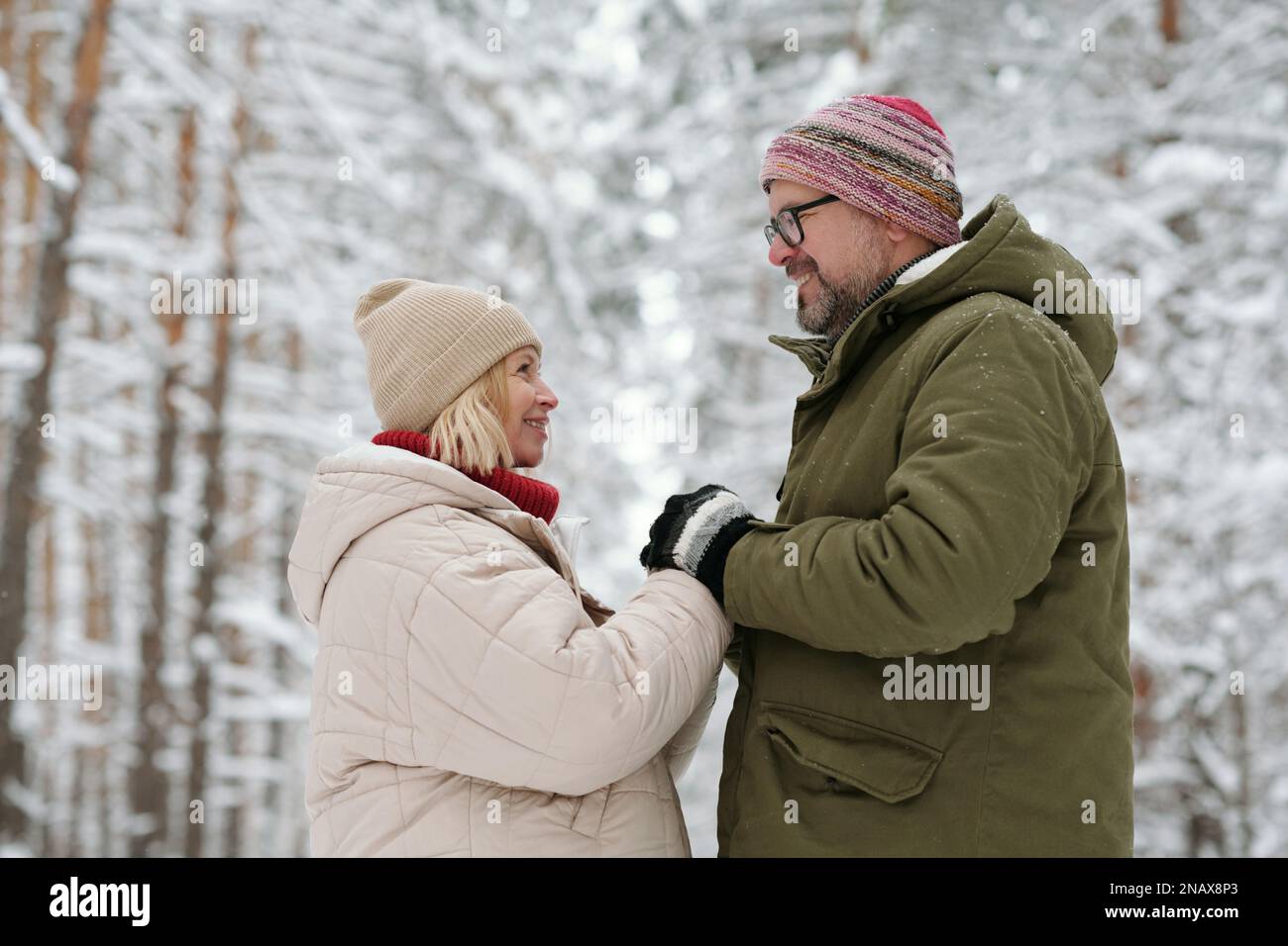 Reifes heterosexuelles Paar, das sich anschaut, während es im Winterwald inmitten von schneebedeckten hohen Kiefernwäldern an den Händen hält Stockfoto