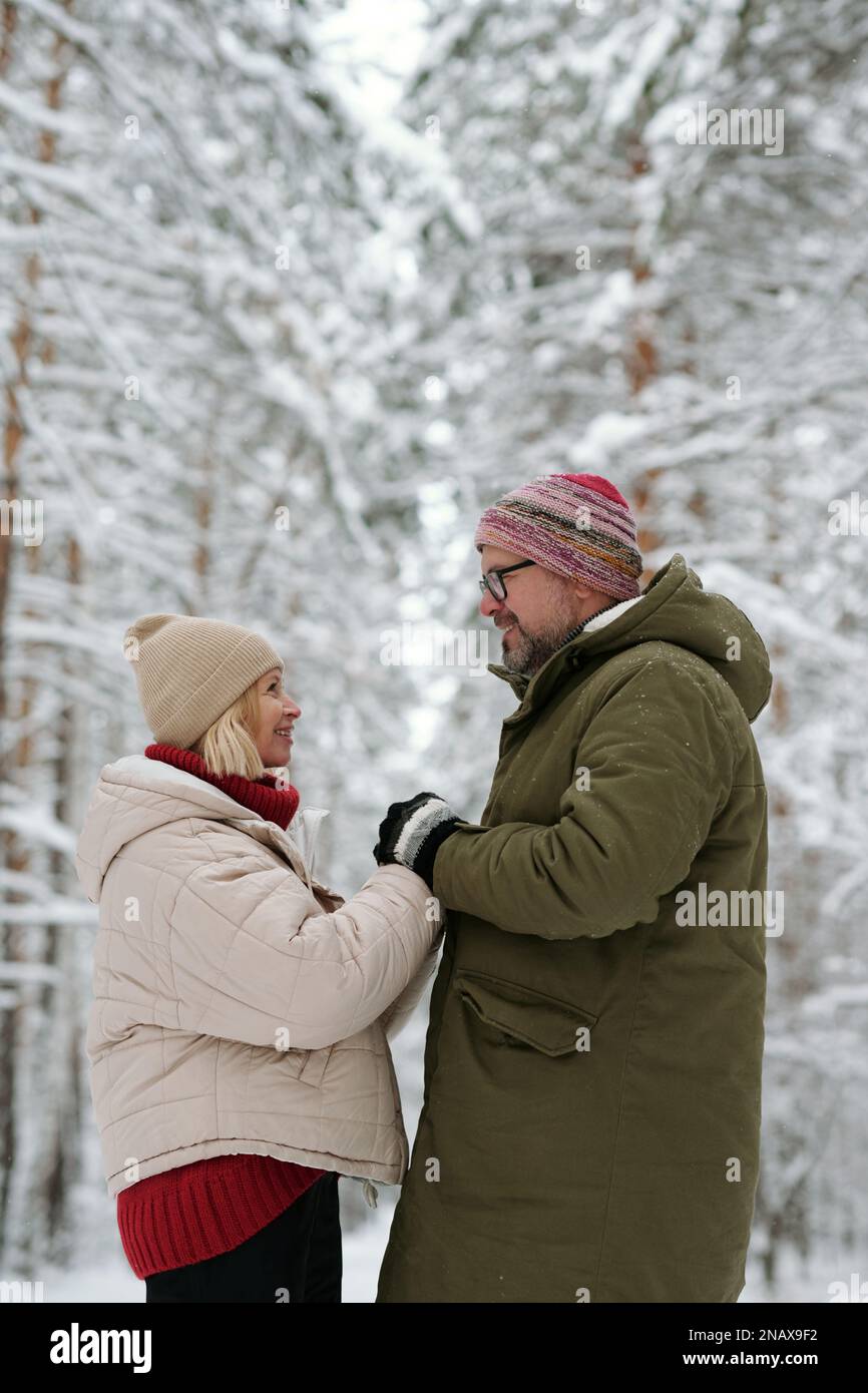 Lächelndes Seniorenpaar, das sich an den Händen festhält und sich anschaut, während es vor der Kamera gegen schneebedeckte Kiefernwälder steht Stockfoto