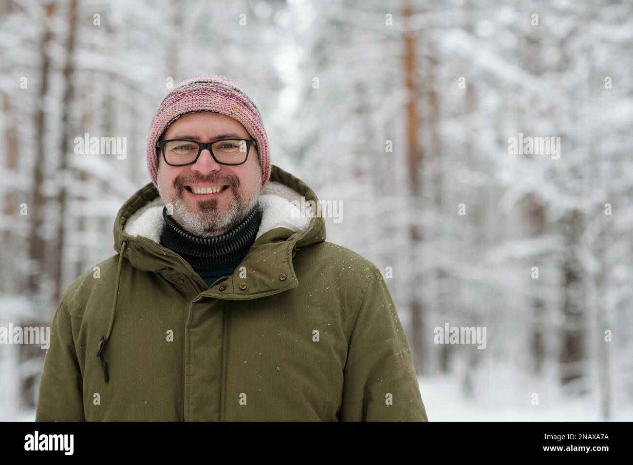 Glücklicher erwachsener Mann in warmer Winterjacke mit Kapuze, Brille und Strickmütze, der im Wald vor der Kamera steht Stockfoto