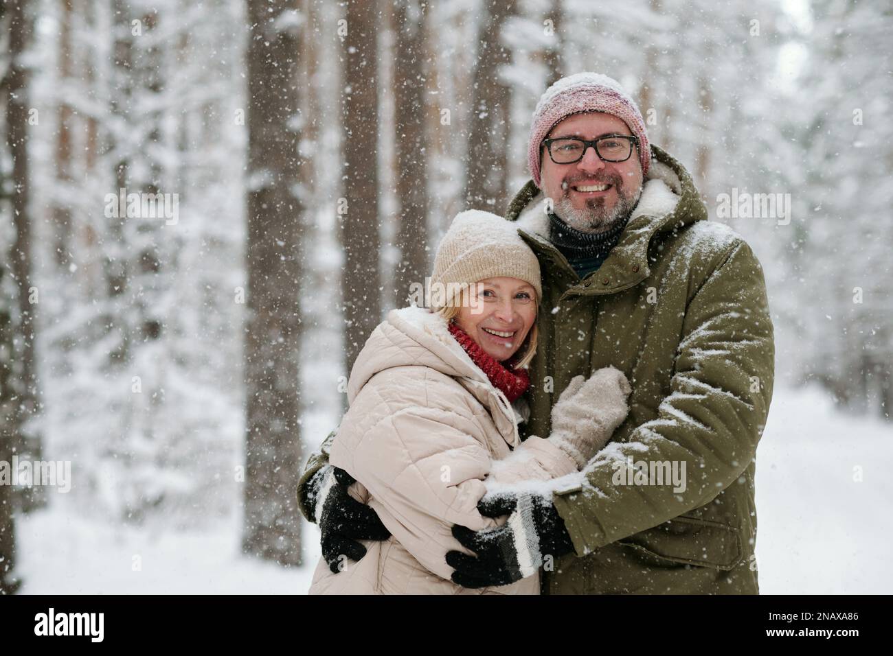 Fröhliches, reifes Paar in Winterbekleidung, das während des Schneefalls in einem schneebedeckten Wald in die Kamera schaut, während ein Mann seine Frau umarmt Stockfoto