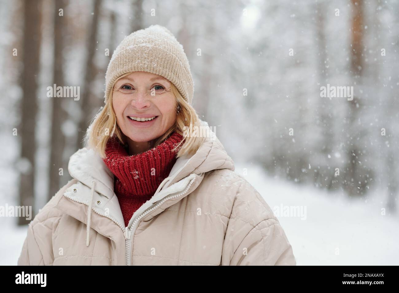 Glückliche, reife blonde Frau in gestrickter Beanie aus Wolle, rotem Pullover und warmer Winterjacke, die im Wald während des Schneefalls auf die Kamera schaut Stockfoto