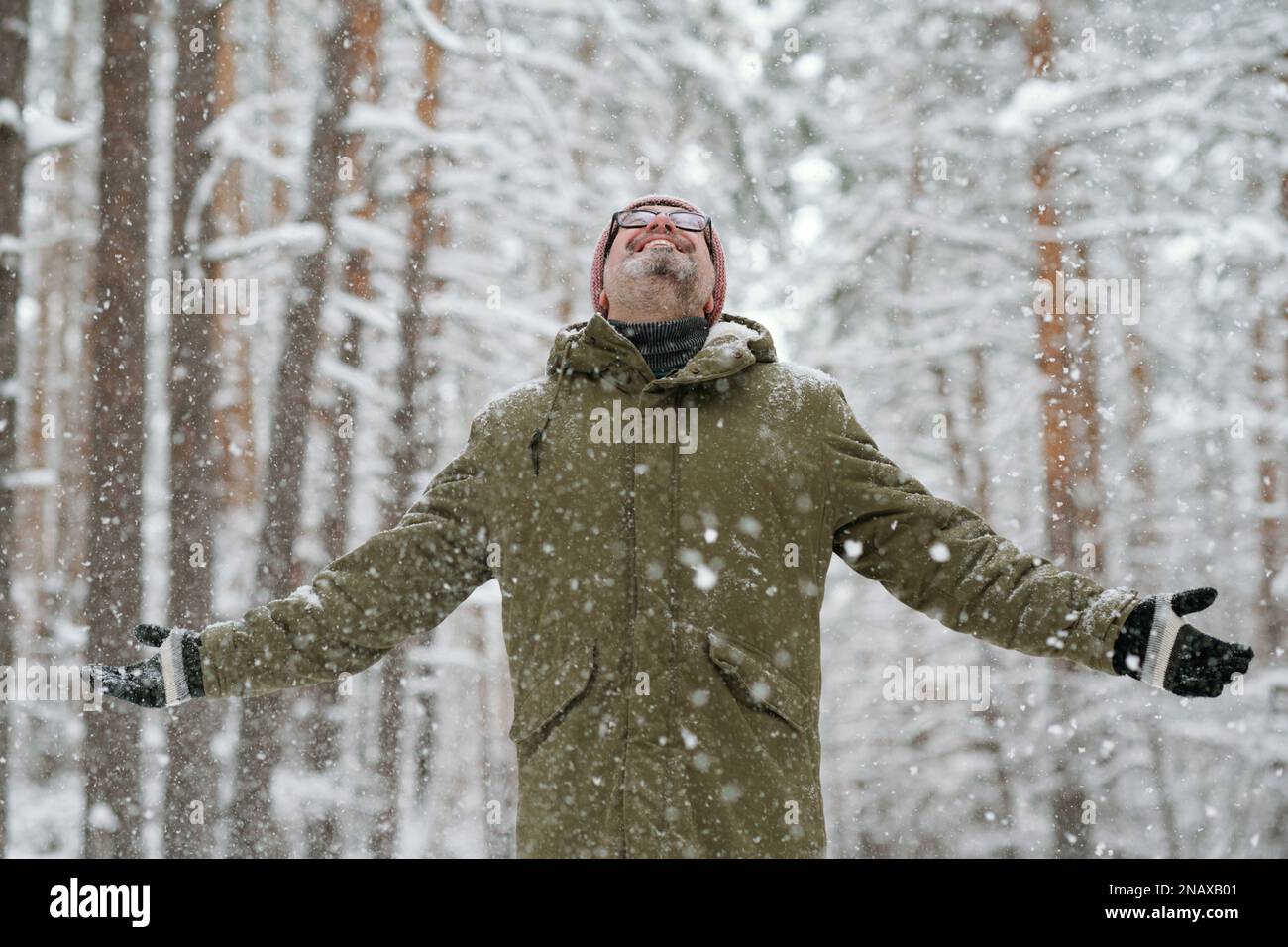 Ein fröhlicher Seniorenmann in Winterjacke und Mütze hält die Arme ausgestreckt, während er nach oben blickt und den Schneefall genießt Stockfoto