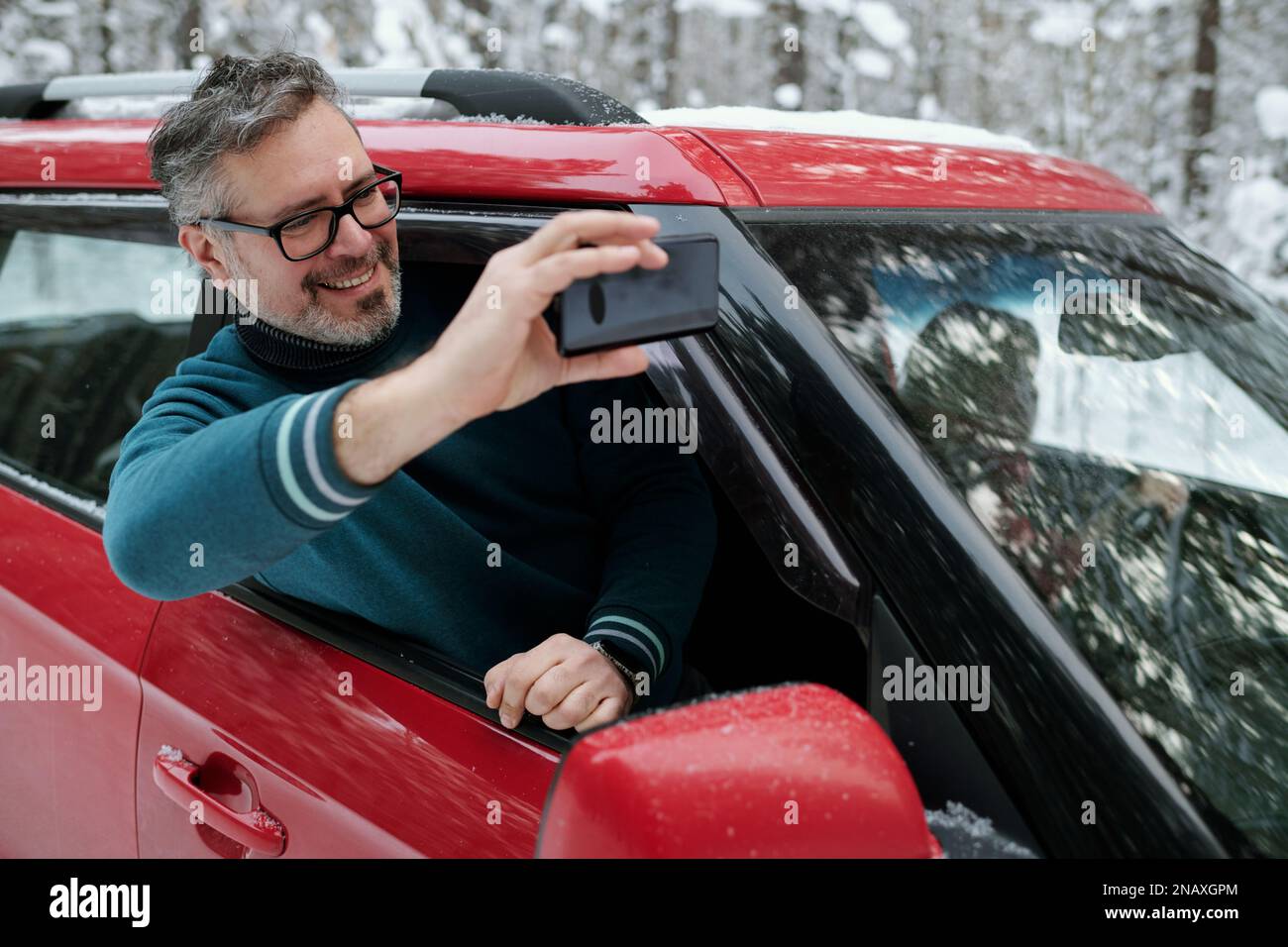 Fröhlicher, reifer Mann mit Mobiltelefon, der Selfie macht, während er während einer Winterreise in natürlicher Umgebung aus dem Autofenster steigt Stockfoto