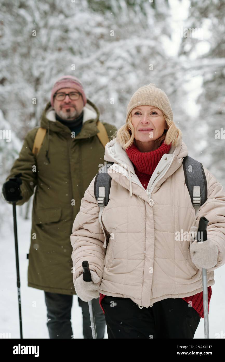 Aktive ältere blonde Frau und ihr Mann mit Wanderstöcken und Rucksäcken, die mit schneebedeckten Bäumen im Park oder Wald spazieren Stockfoto