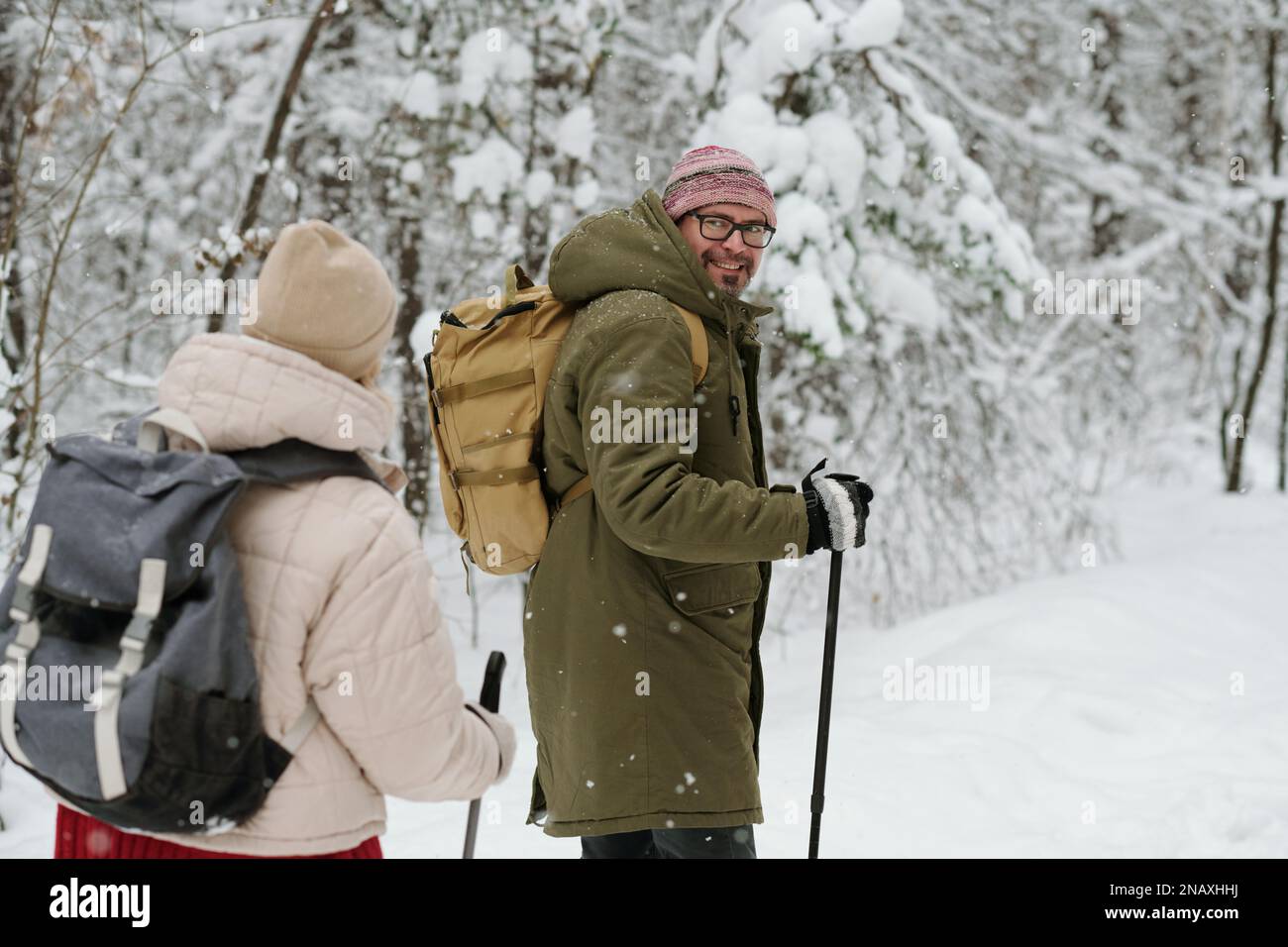 Glücklicher, reifer Mann mit Rucksack und Wanderstöcken, der seine Frau ansieht, während beide auf der schneebedeckten Straße im Wald spazieren Stockfoto