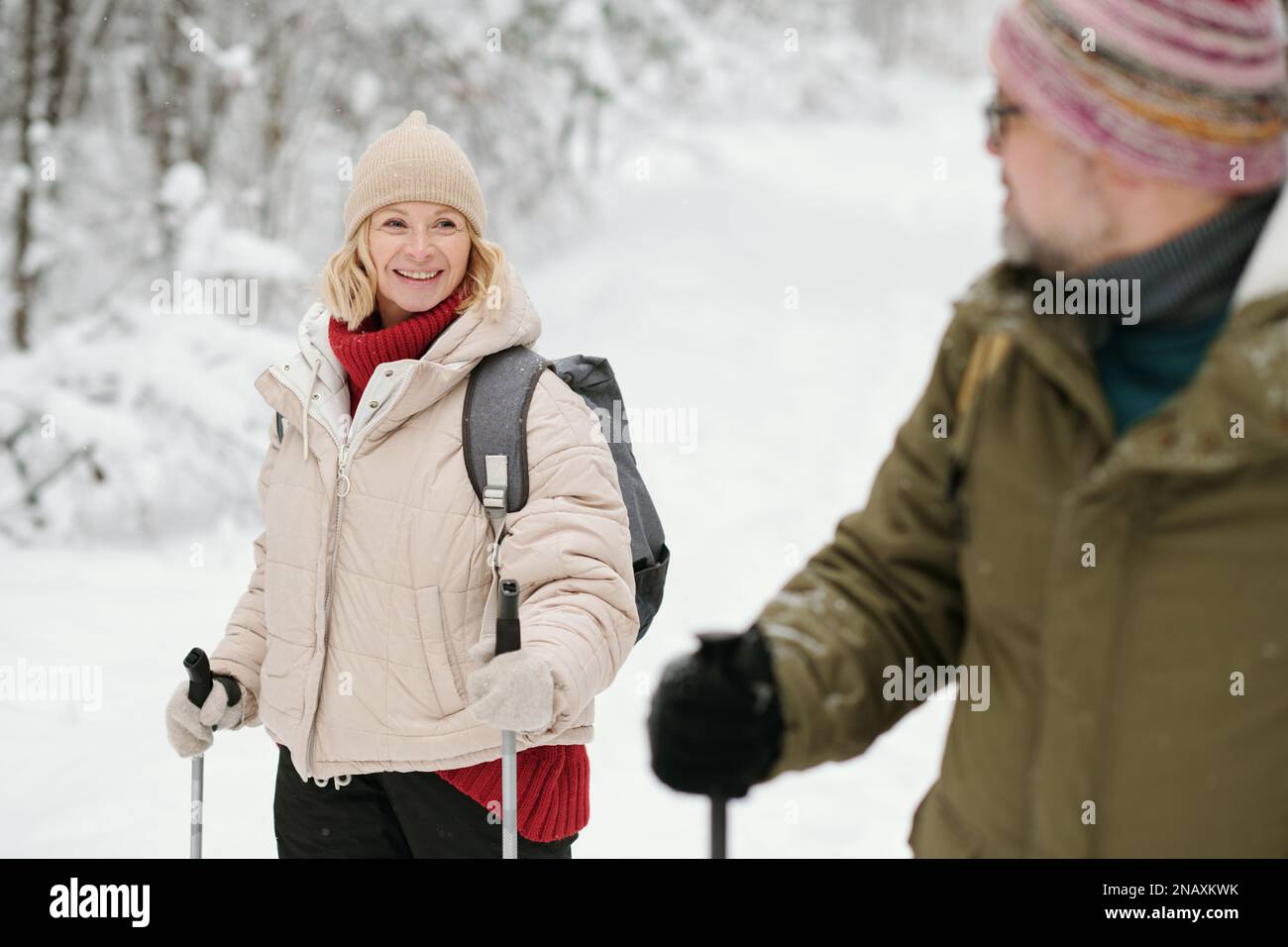 Eine reife lächelnde Frau in warmer Winterbekleidung, die ihren Mann während des Spaziergangs mit Wanderstöcken im Wald oder im schneebedeckten Park ansieht Stockfoto