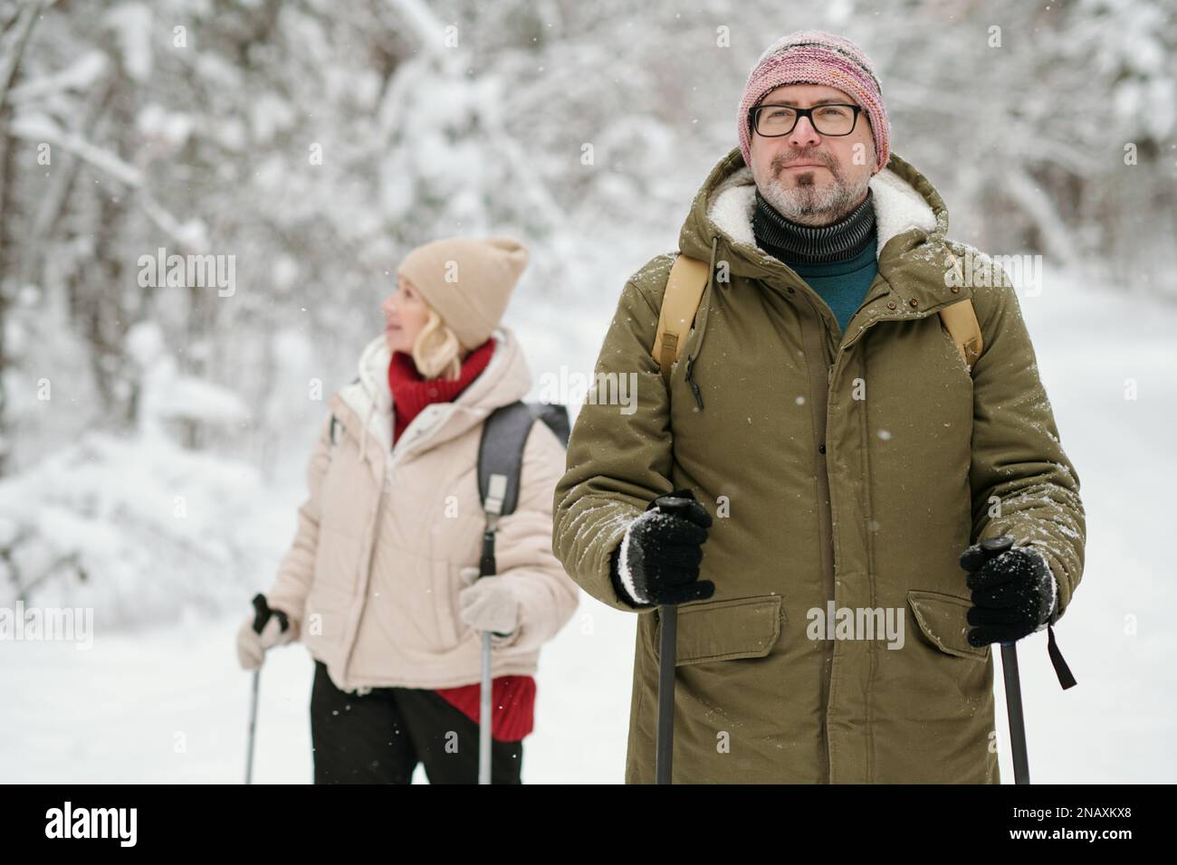 Reifer Mann in Winterbekleidung, der mit Wanderstöcken im schneebedeckten Wald spaziert, während seine Frau im Hintergrund steht Stockfoto