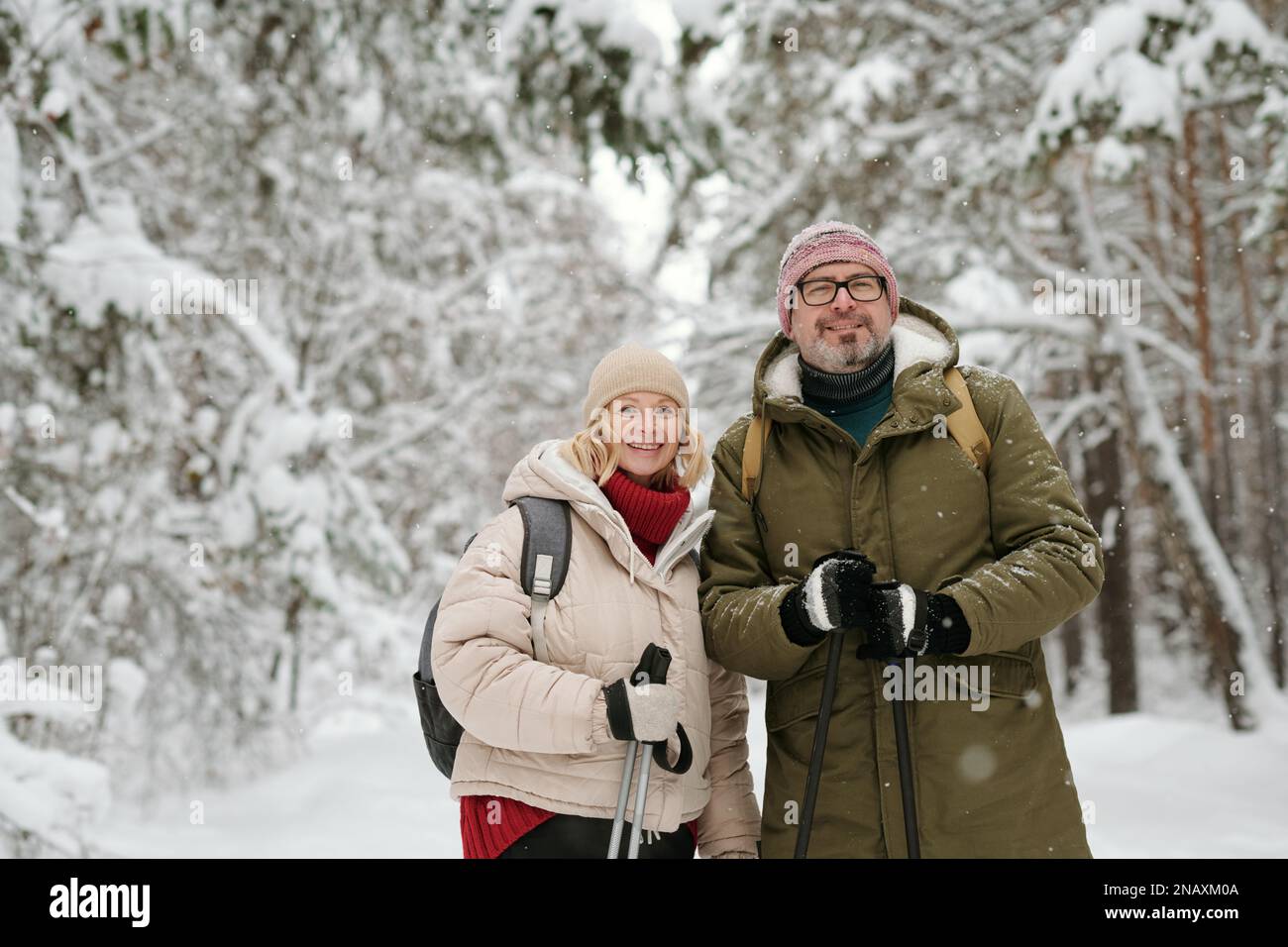 Älterer, lächelnder Ehemann und Ehefrau mit Trekkingstöcken, die während des Spaziergangs in einem schneebedeckten Wald stehen und in die Kamera schauen Stockfoto