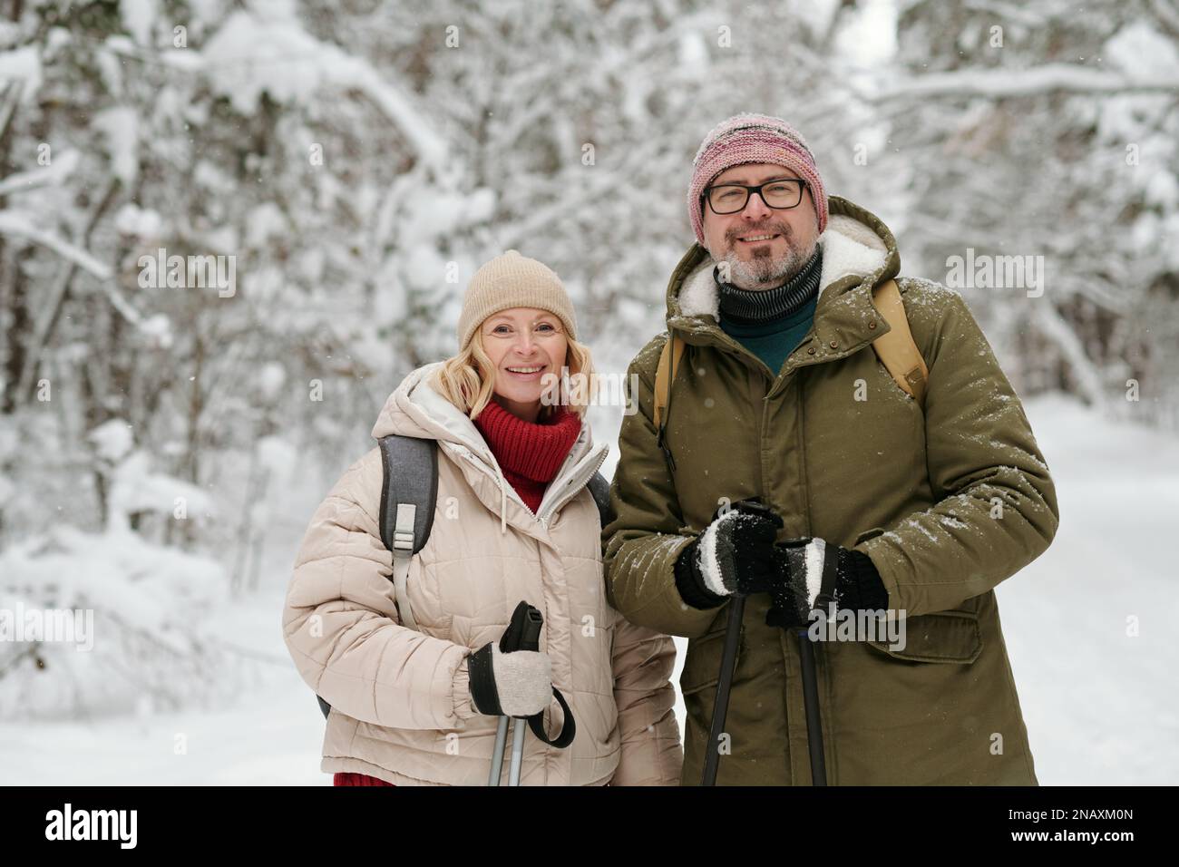 Fröhliches Seniorenpaar mit Wanderstöcken, die auf die Kamera schauen, während Sie am Wochenende im Winterwald spazieren gehen, der mit Schnee bedeckt ist Stockfoto