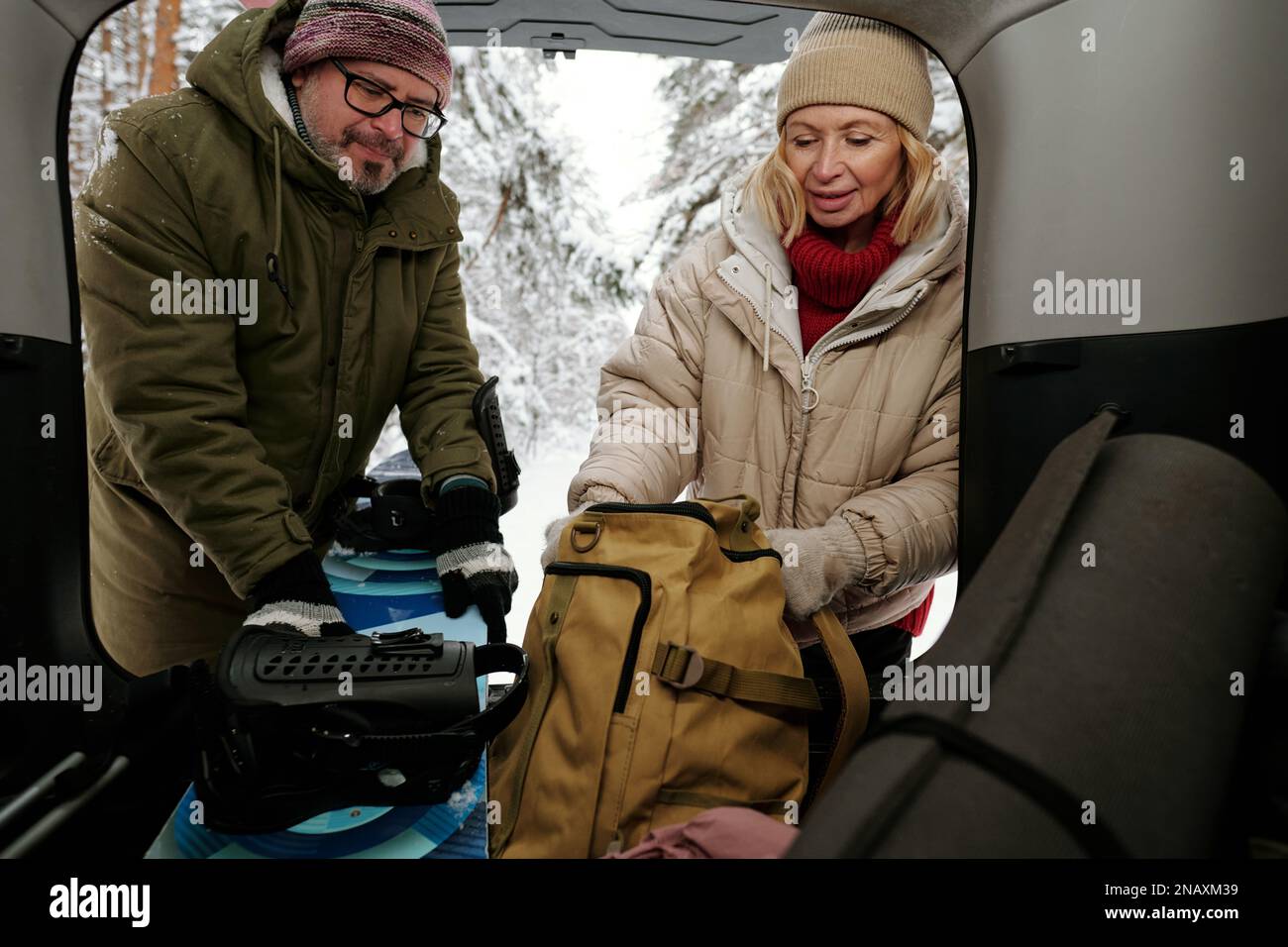 Zwei reife Touristen in Winterkleidung, die das Snowboard aus dem Kofferraum nehmen, während eine Frau ihrem Mann hilft und den Rucksack mit Essen in der Hand hält Stockfoto