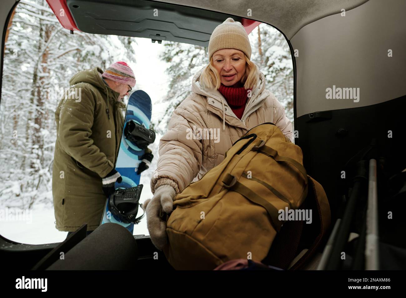 Reife Frau in Winterjacke und warmer Beanie, die den Rucksack in den Kofferraum steckt, während ihr Mann sich auf das Snowboarden im Wald vorbereitet Stockfoto