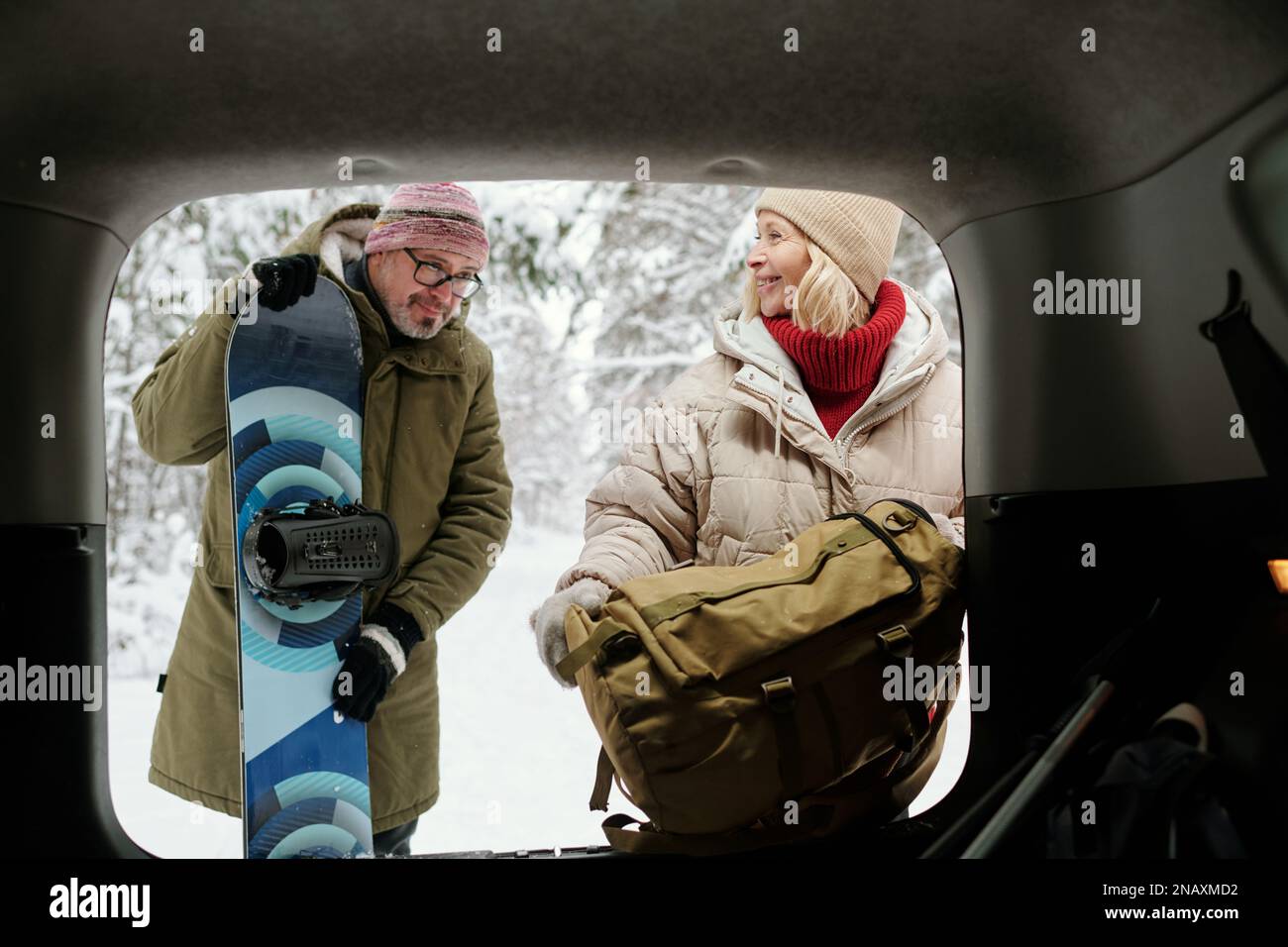 Glückliche blonde Frau mit Rucksack, die neben ihrem Mann mit Snowboard am offenen Kofferraum stand, während beide ihre Winterreise genossen Stockfoto