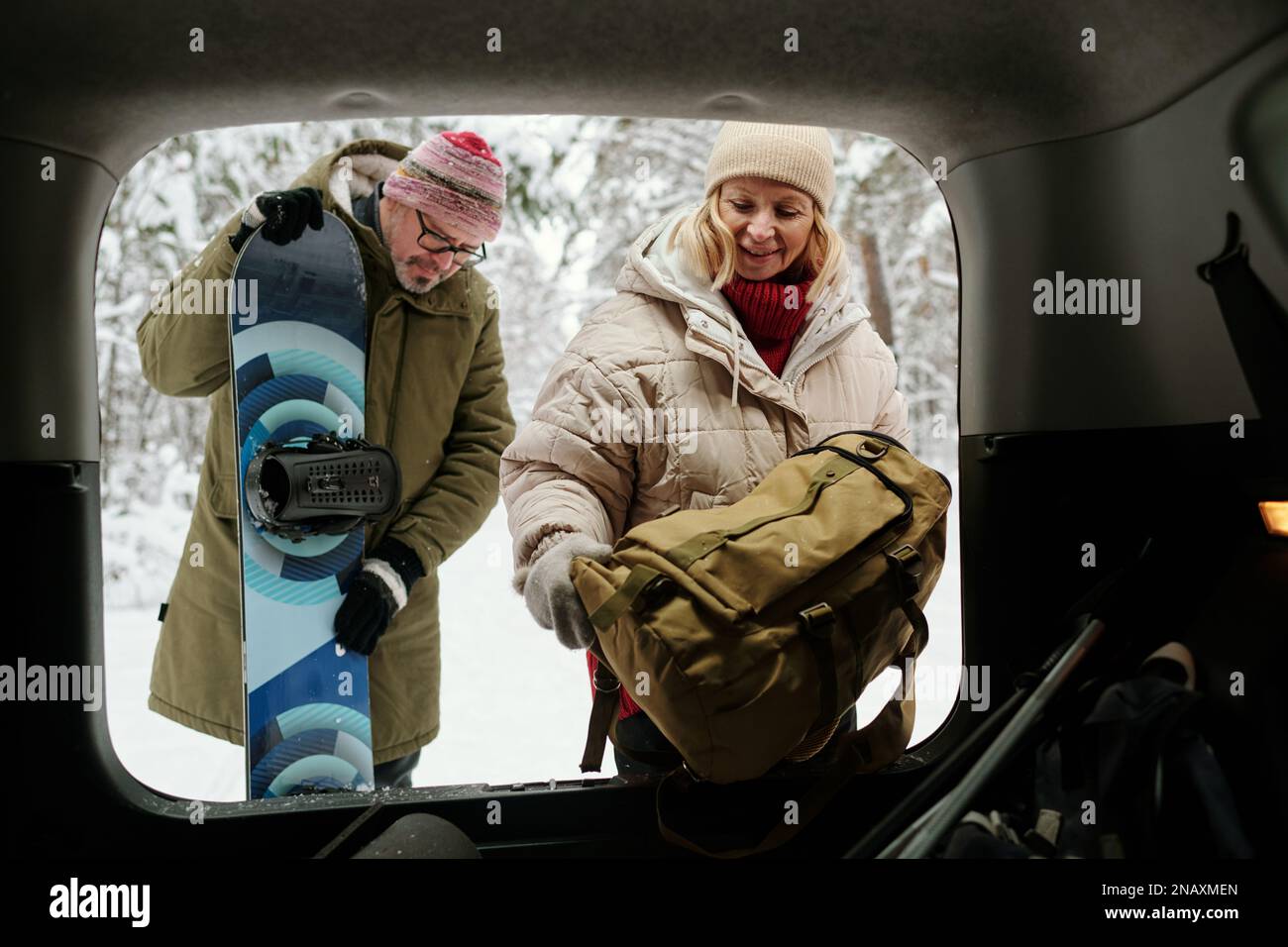 Seniorin legt Rucksack in den Kofferraum, während ihr Mann mit Snowboard während ihres Wochenendausflugs neben ihr steht Stockfoto