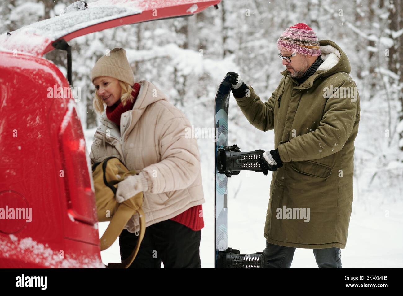 Aktiver Senior-Mann mit Snowboard hinter seiner Frau, der während seiner Reise in den Winterwald einen Rucksack in den Kofferraum steckte Stockfoto