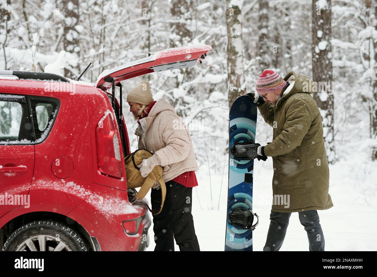 Seitenansicht eines aktiven erwachsenen Ehepaars in Winterbekleidung, das sein Gepäck aus dem Kofferraum des Fließhecks nimmt, während ein älterer Mann auf das Snowboard schaut Stockfoto