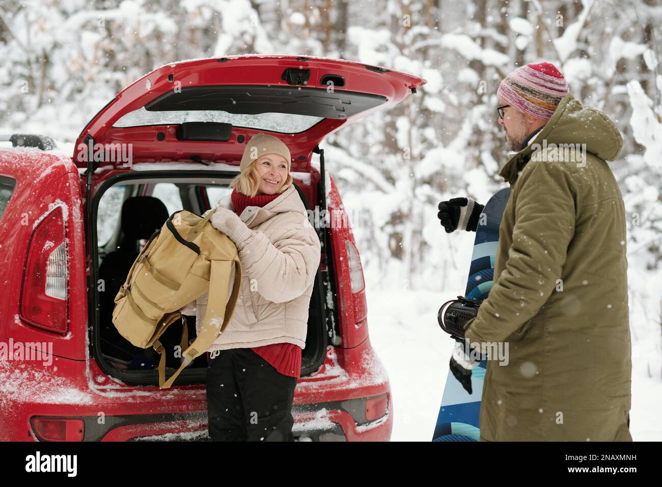 Glückliche ältere Frau mit Rucksack, die ihren Mann mit Snowboard ansieht, während sie Gepäck aus dem Kofferraum nimmt Stockfoto
