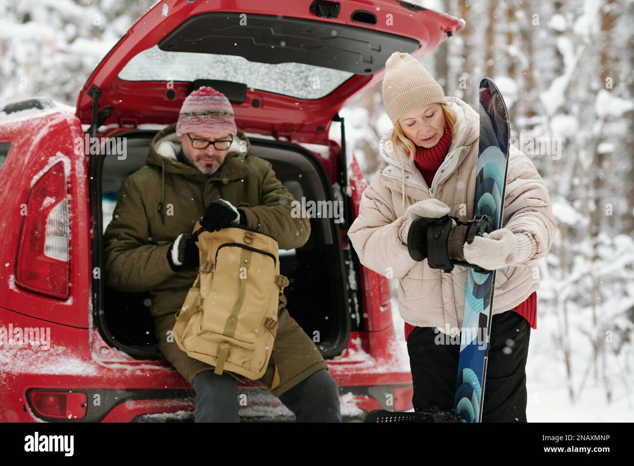 Reife blonde Frau, die während des Winterurlaubs Snowboard schaut, während ihr Mann im Kofferraum sitzt und den Reißverschluss aufmacht Stockfoto