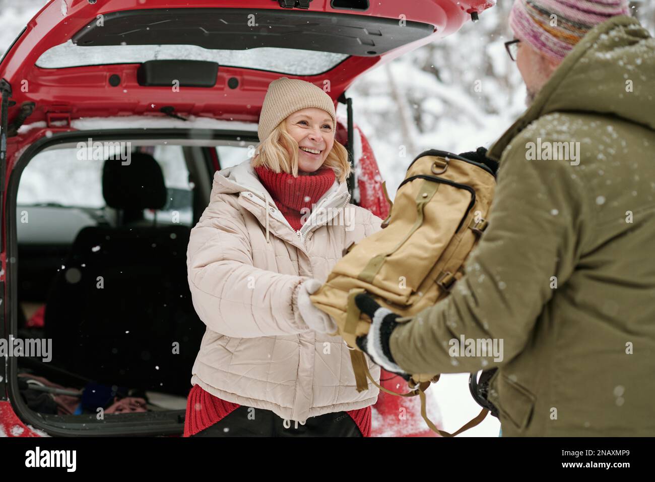 Reife, fröhliche Frau in warmer Winterjacke und Mütze, die ihren Rucksack mit Proviant an ihren Mann weitergibt, während sie am Auto steht Stockfoto