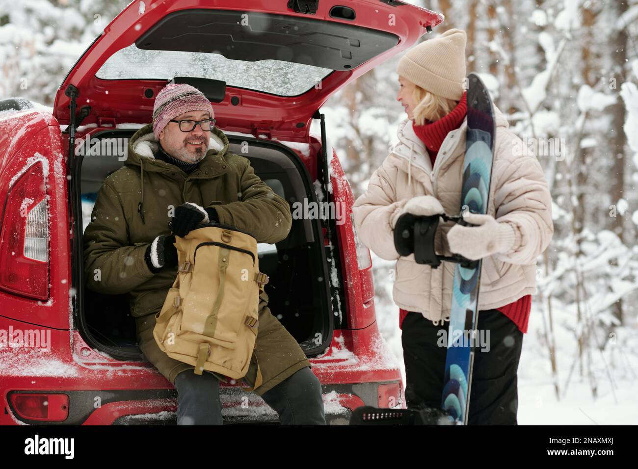 Reife, aktive Frau mit Snowboard, die ihren Mann beim Ausziehen des Rucksacks ansieht, während er während der Winterwanderung im Kofferraum des Sattelautos sitzt Stockfoto