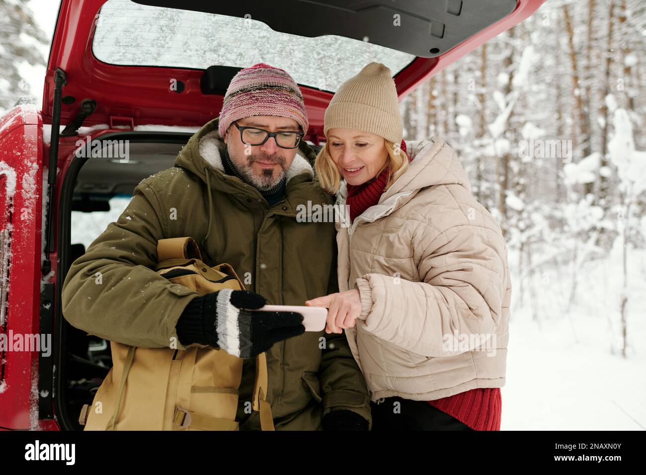 Glückliche, reife Frau, die auf den Bildschirm des Smartphones zeigt, das ihr Mann in der Hand hält, während sie ihm während der Winterreise ein neugieriges Foto zeigt Stockfoto