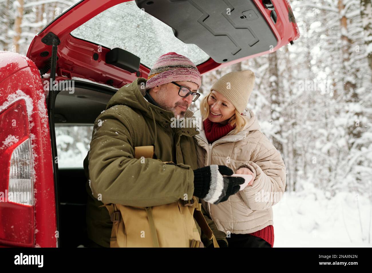 Ein reifer Mann, der sich neue Reisefotos auf dem Smartphone-Bildschirm ansieht, während er im Winterwald neben seiner Frau am Kofferraum des Cabriolet steht Stockfoto