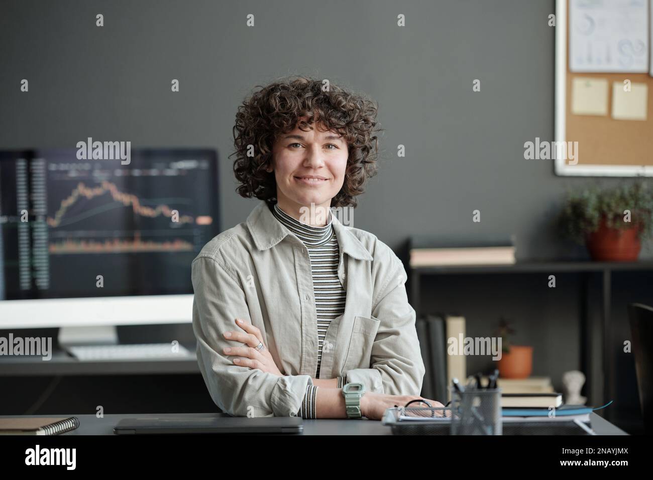 Junge, erfolgreiche Finanzanalytikerin, die am Arbeitsplatz im Büro sitzt und vor die Kamera schaut, um die Grafikdaten auf dem Computerbildschirm abzugleichen Stockfoto