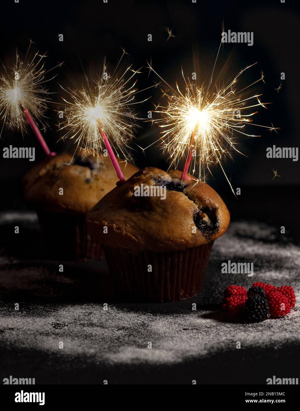 Feiern Sie die süßen Momente des Lebens mit diesen köstlichen Cranberry-Muffins, geschmückt mit glitzernden Kerzen vor schwarzem Hintergrund Stockfoto