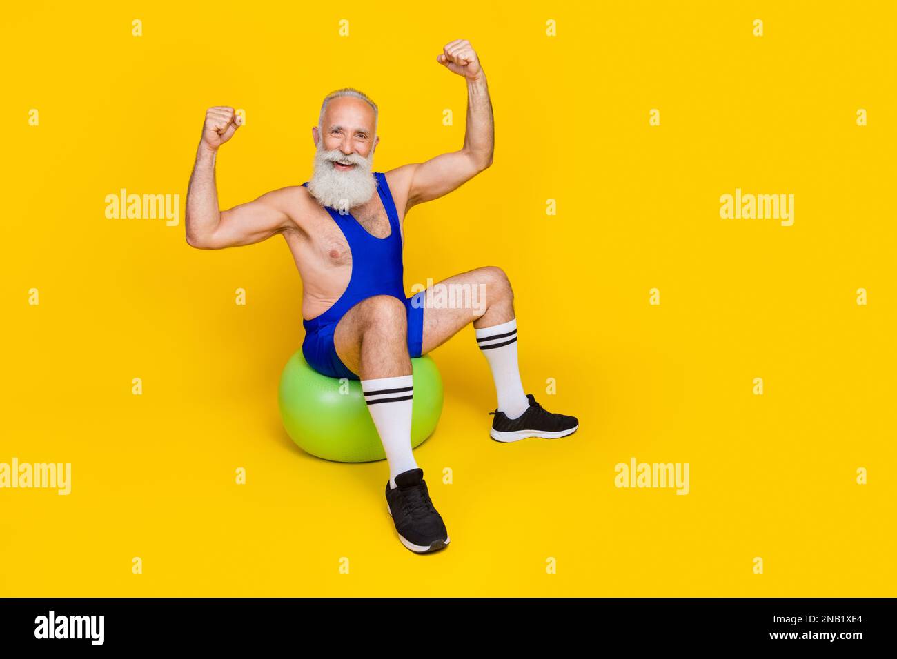 Langes Foto des alten lustigen Großvaters mit Bart Sitzkleidung blaue Sportbekleidung, auf dem die Hände hoch sind starke Muskeln isoliert auf gelber Farbe Stockfoto