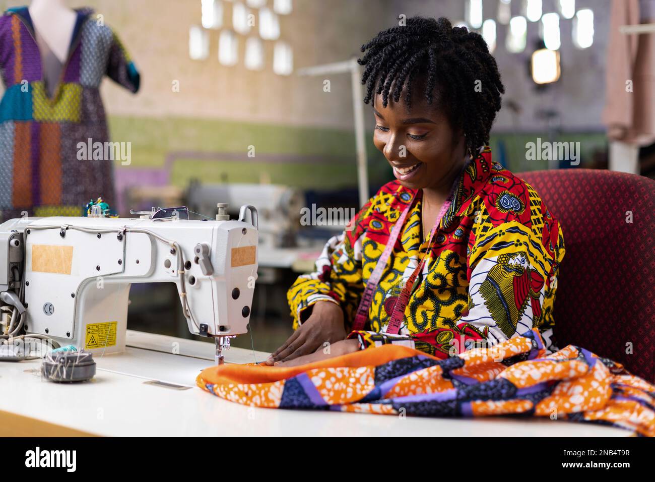 Eine afrikanische Modeschneiderin trägt ein ankara-Kleid mit locs-Haaren und näht ein Outfit in ihrem Dressmaking-Studio mit einer elektrischen Maschine. Stockfoto