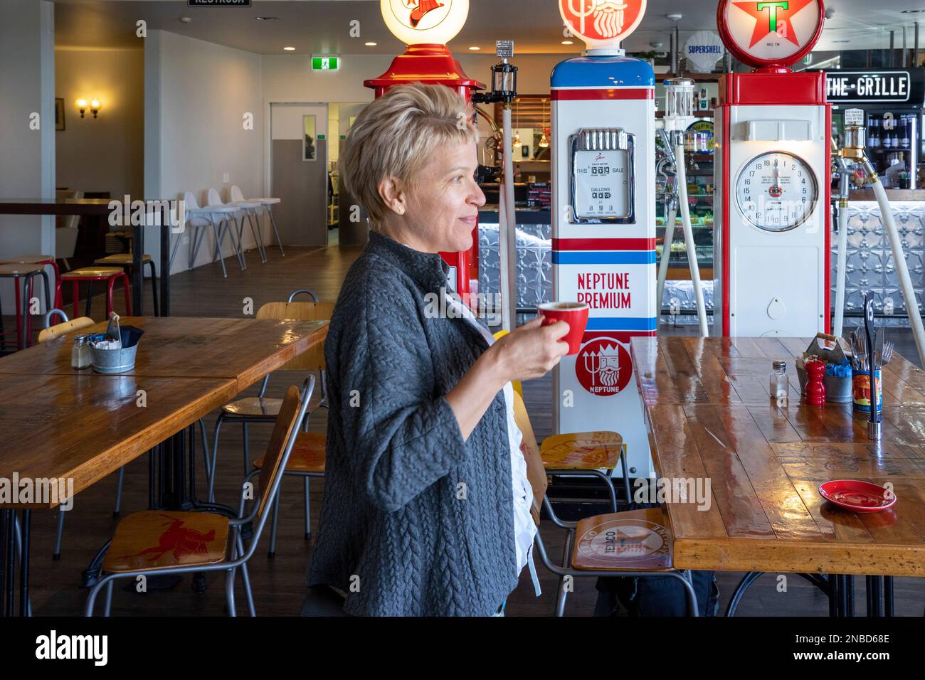 Blonde weiße Frauen, die Kaffee aus dem roten Becher im alten Tankstellencafé trinken, mit den Logos und Schildern verschiedener Benzinhersteller. Stockfoto