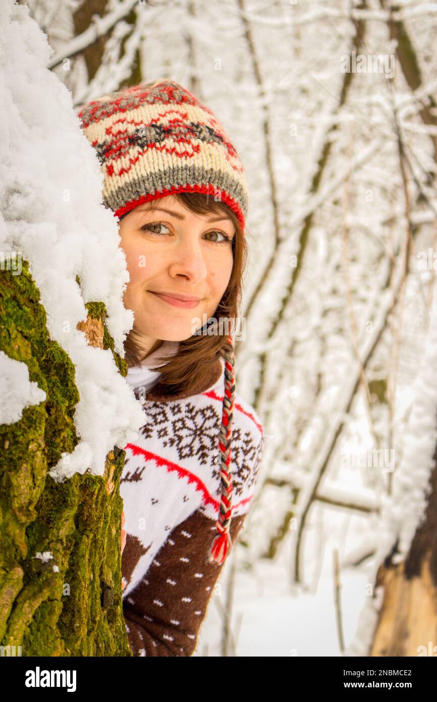 Lächelndes junges Mädchen in bequemer Strickkleidung im verschneiten Winterwald. Winterwetter- und Bekleidungskonzept. Glückliche Frau in Hirschpullover und nepalesischer Mütze. Stockfoto