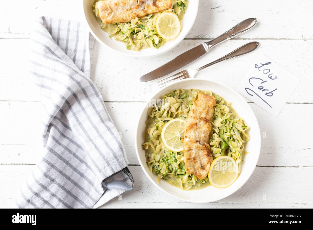 Kohlenhydratarmes Abendessen oder Mittagessen mit gebratenem Fischfilet in der Pfanne und cremigen Zucchini-Nudeln. Serviert auf Tellern auf weißem Hintergrund mit Besteck Stockfoto