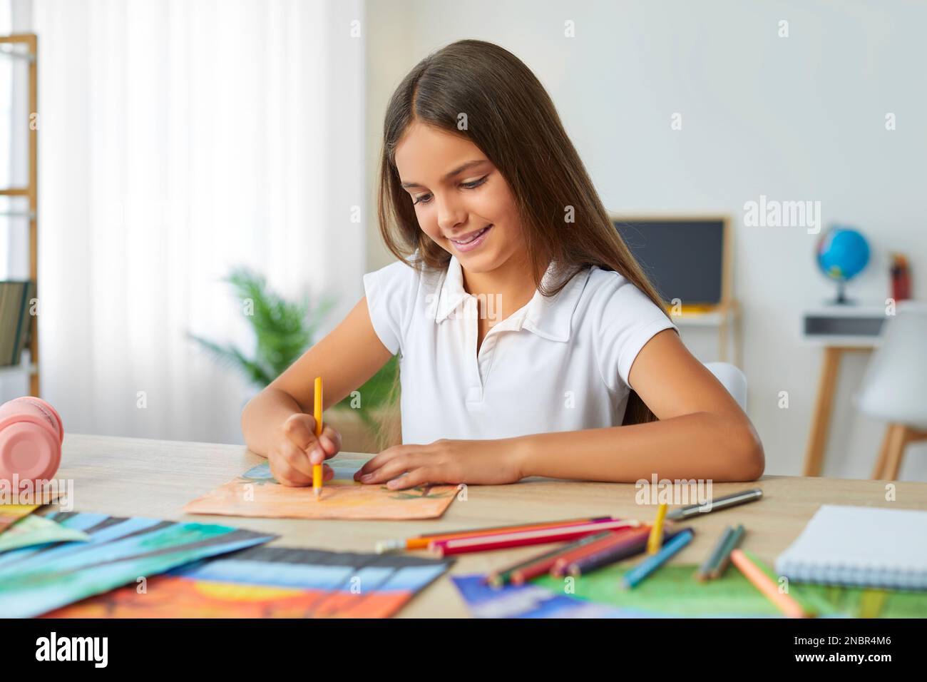 Ein lächelndes, süßes Mädchen malt ein Bild mit Interesse und sitzt zu Hause am Tisch. Stockfoto