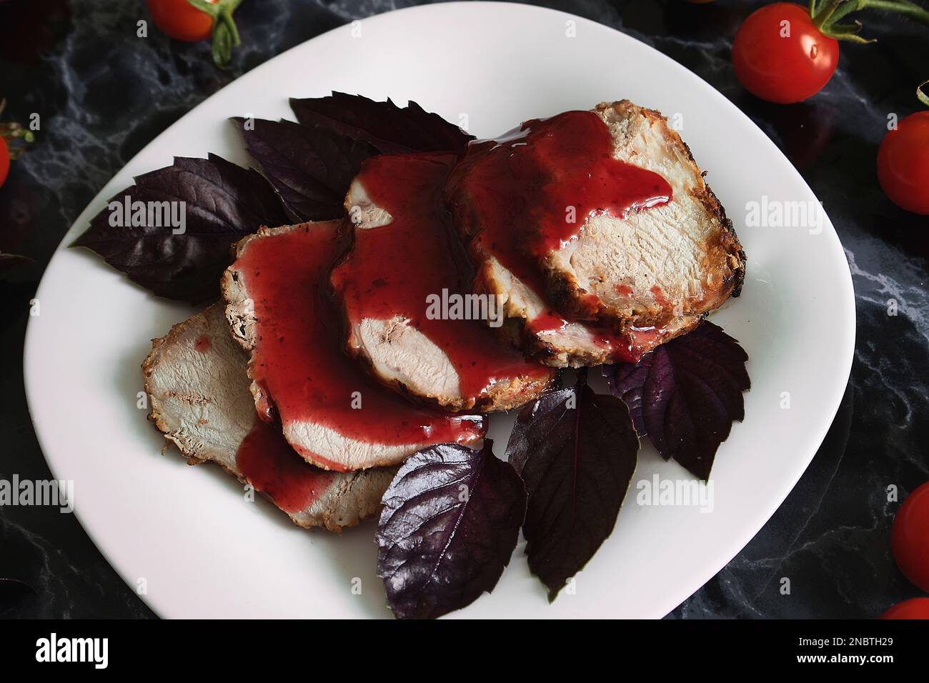 Saftiges Steak, gut gebratenes Schweinefleisch mit roter Sauce und Basilikumblättern auf einem weißen Teller über einem schwarzen Marmortisch. Kirschtomaten und Basilikum-Zweige auf einer mär Stockfoto