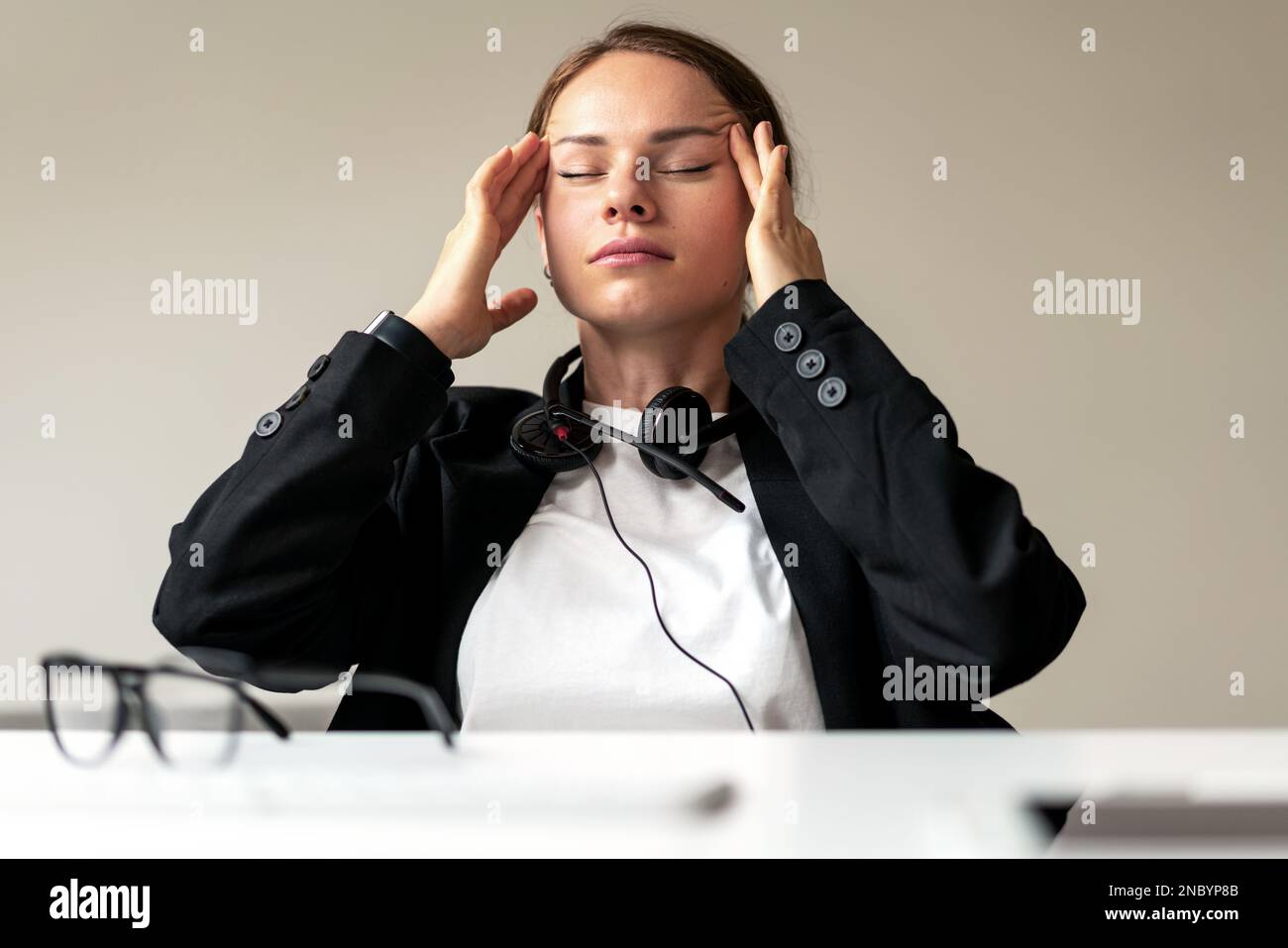 Müde Geschäftsfrau, die mit geschlossenen Augen am Schreibtisch sitzt und den Tempelbereich mit den Händen reibt. Stockfoto
