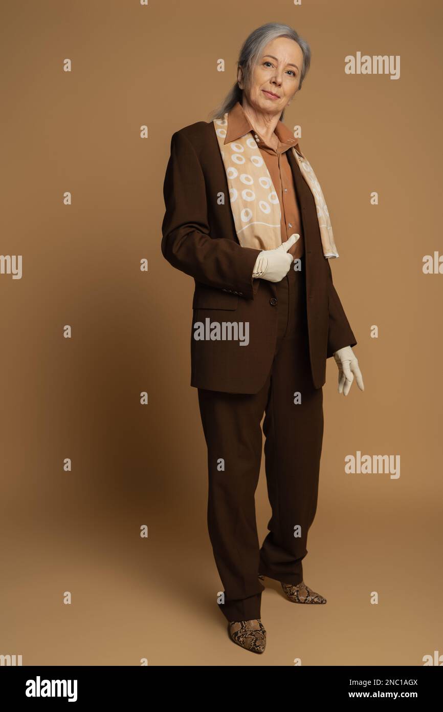 Die ganze Länge einer älteren Frau in brauner formeller Kleidung und weiße Handschuhe, die wie ein Schild in Beige aussehen Stockfoto