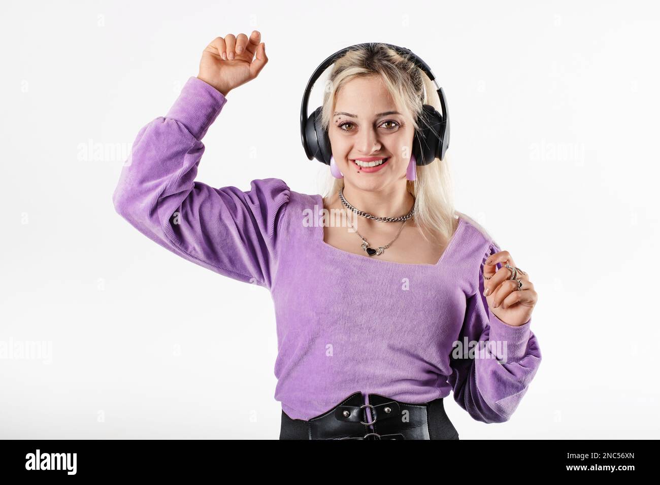 Die fröhliche blonde Frau trägt eine lila gerippte Bluse, isoliert auf weißem Hintergrund, lächelt in die Kamera und tanzt über Kopfhörer zur Musik. Stockfoto