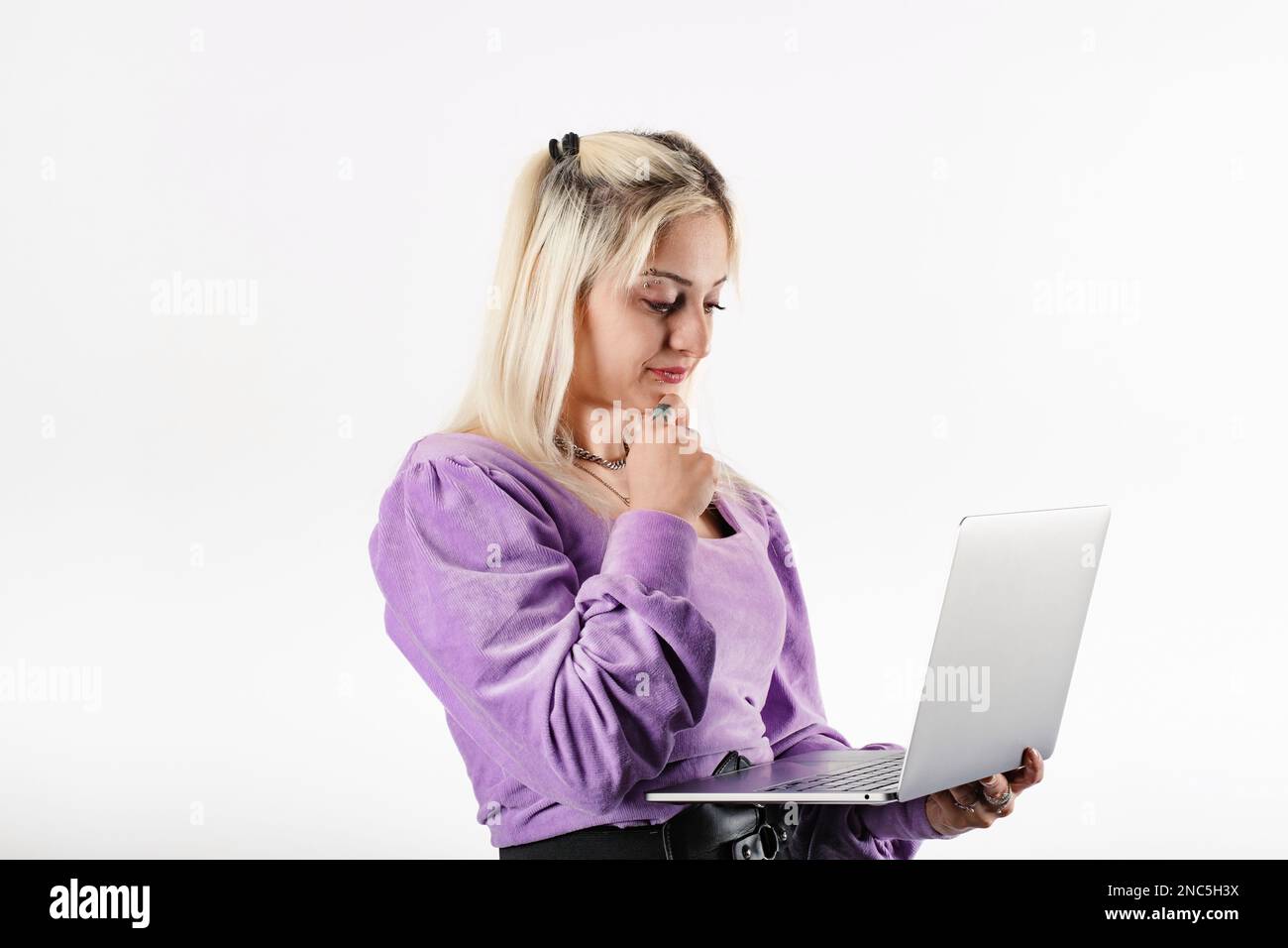 Blonde Frau, die isoliert auf weißem Hintergrund steht und den Laptop mit einem selbstbewussten Gesichtsausdruck hält, das Kinn berührt und auf den Computer schaut. Lesen a b Stockfoto