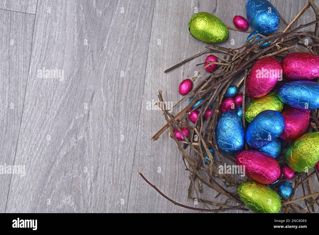 In Folie verpackte mehrfarbige ostereier in Rosa, Grün, Blau und Gelb in einem natürlichen Nest aus Stöcken und Zweigen auf grauem Holzhintergrund. Stockfoto