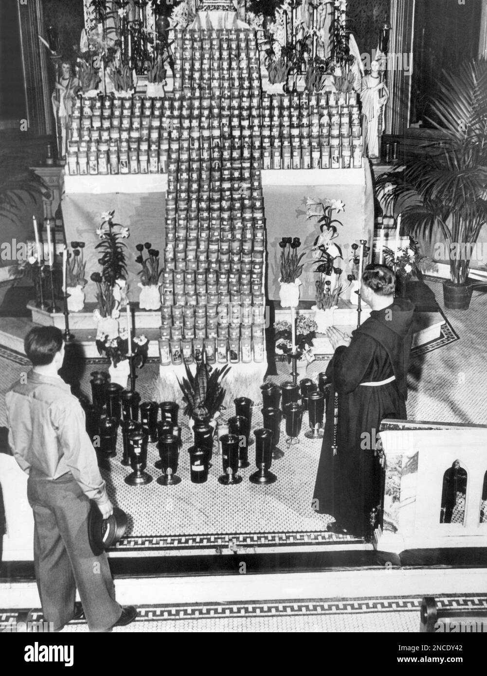 A soldier and a priest offer a prayer for servicemen serving overseas as they stand before the alter in San Sebastian’s Church in New York on March 31, 1945, where 450 candles in votive lamps have been arranged in the form of an Easter cross. Each candle is for a serviceman serving abroad, and each container bears the flag and a holy picture. The soldier is unable to kneel during the prayer because of a leg injury received in France. (AP Photo) Stockfoto
