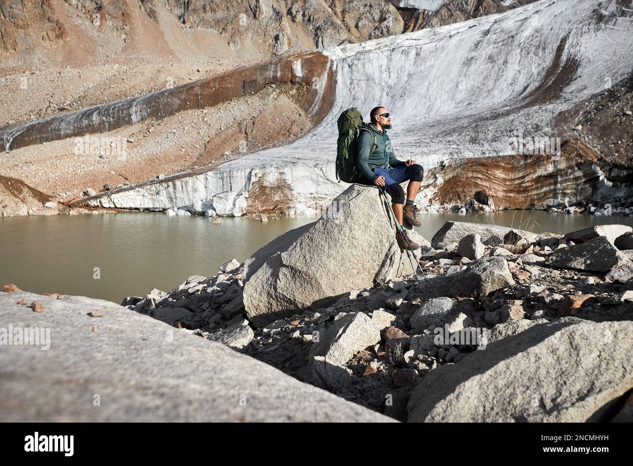 Bärtiger Wandertouristin mit großem Rucksack und Wanderstöcken auf dem Felsen in der Nähe des Bergsees und des Eisgletschers in Kasachstan. Outdoor und Wandern Stockfoto