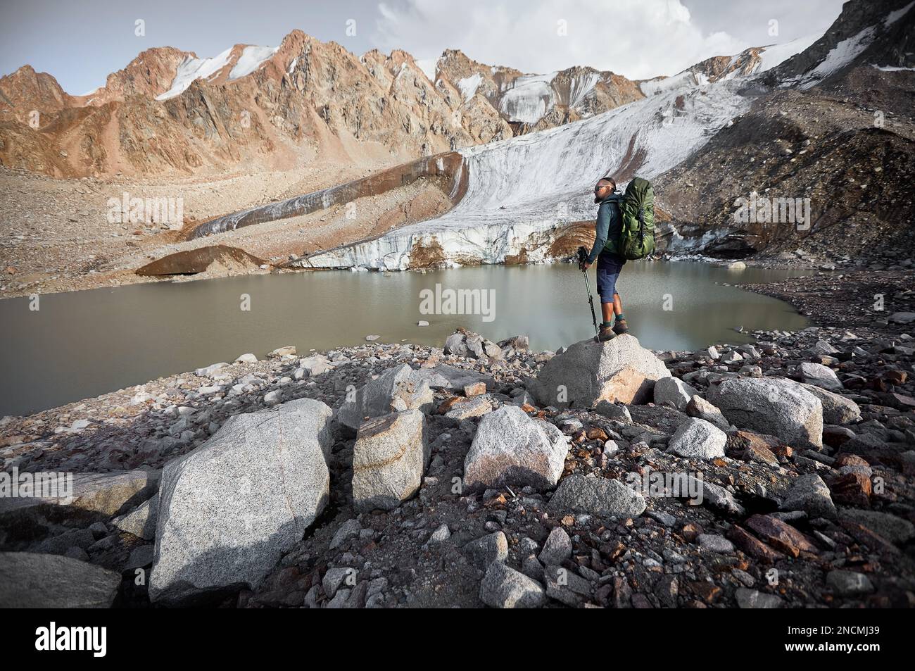 Bärtiger Wanderer mit großem Rucksack und Wanderstöcken auf dem Felsen in der Nähe des Bergsees und des Eisgletschers in Kasachstan. Outdoor- und Trekking-Konz Stockfoto