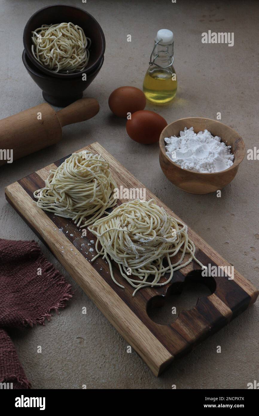 Frische Nudeln auf einem Holztablett mit den Zutaten einer Schüssel Mehl, zwei Eiern und Palmöl Stockfoto