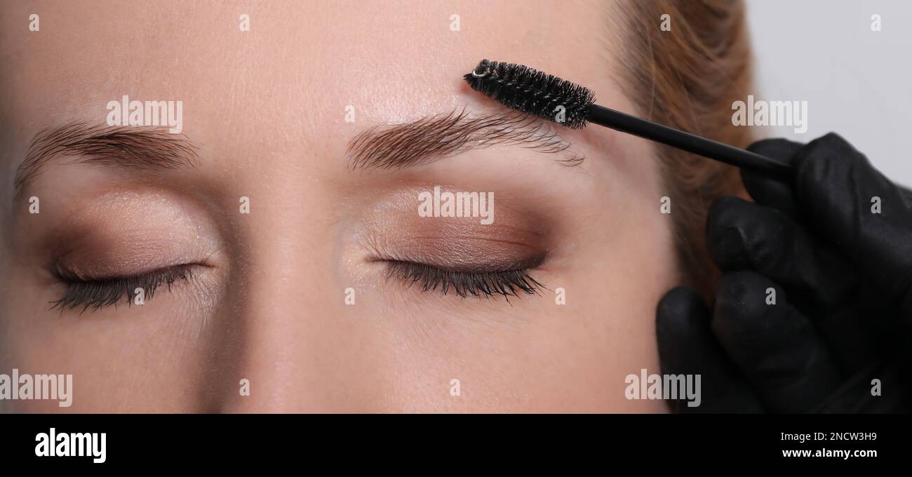 Kosmetikerin putzt die Augenbrauen der Frau, bevor sie auf hellgrauem Hintergrund abfärbt, Nahaufnahme Stockfoto