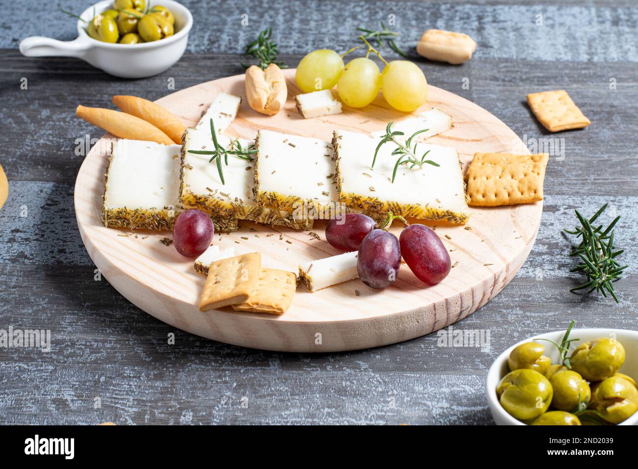 Schmackhafter Käse, serviert auf einem Holztisch mit reifen Trauben und Crackern, dekoriert mit Rosmarin-Zweigen neben Oliven in Schüsseln auf dem Tisch Stockfoto