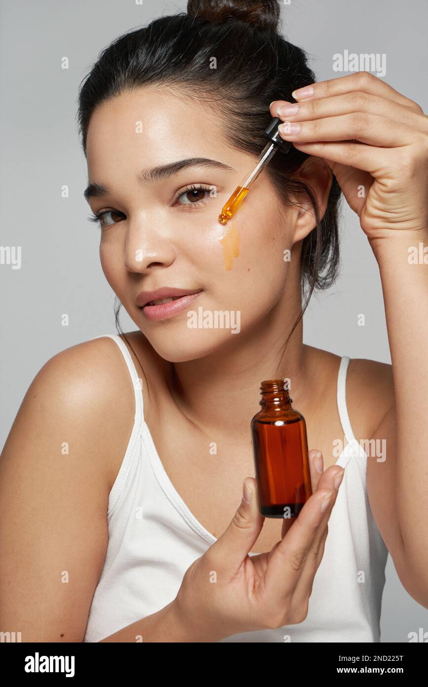 Pensive junge Frau mit weißem Oberteil, die Gesichtsöl aufträgt und dabei vor grauem Hintergrund steht Stockfoto