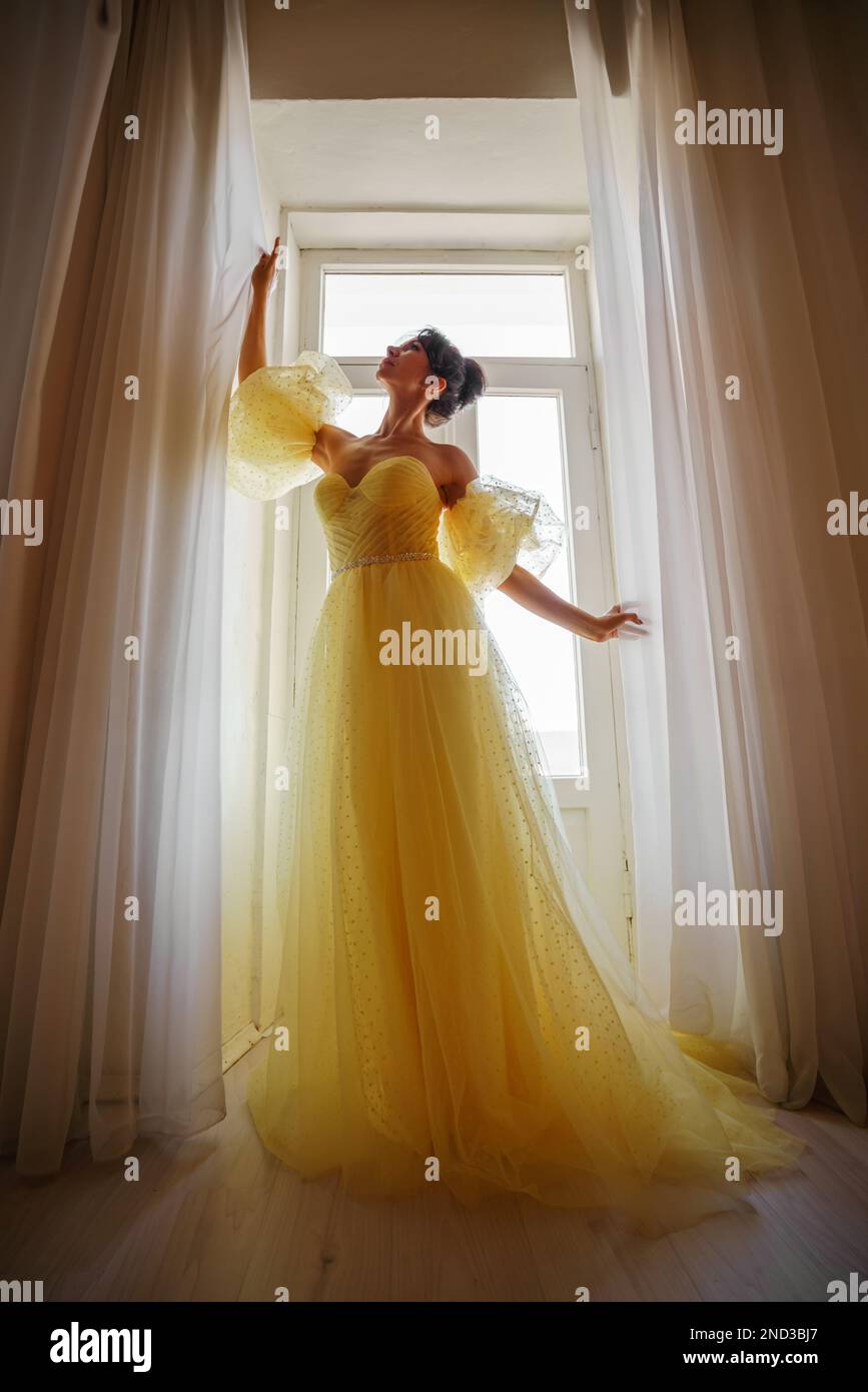 Die Silhouette einer Frau in einem goldenen, luxuriösen Kleid vor dem Hintergrund eines Fensters hält einen Vorhang mit den Händen. Elegante Dame in gelber Seide Stockfoto