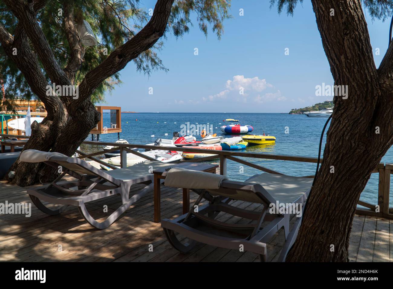 Sonnenliegen im Schatten eines Baumes am Meer. Jet Ski, Wasserschlauch im Wasser, Bodrum Beach Club, Türkei. Selektiver Fokus Stockfoto