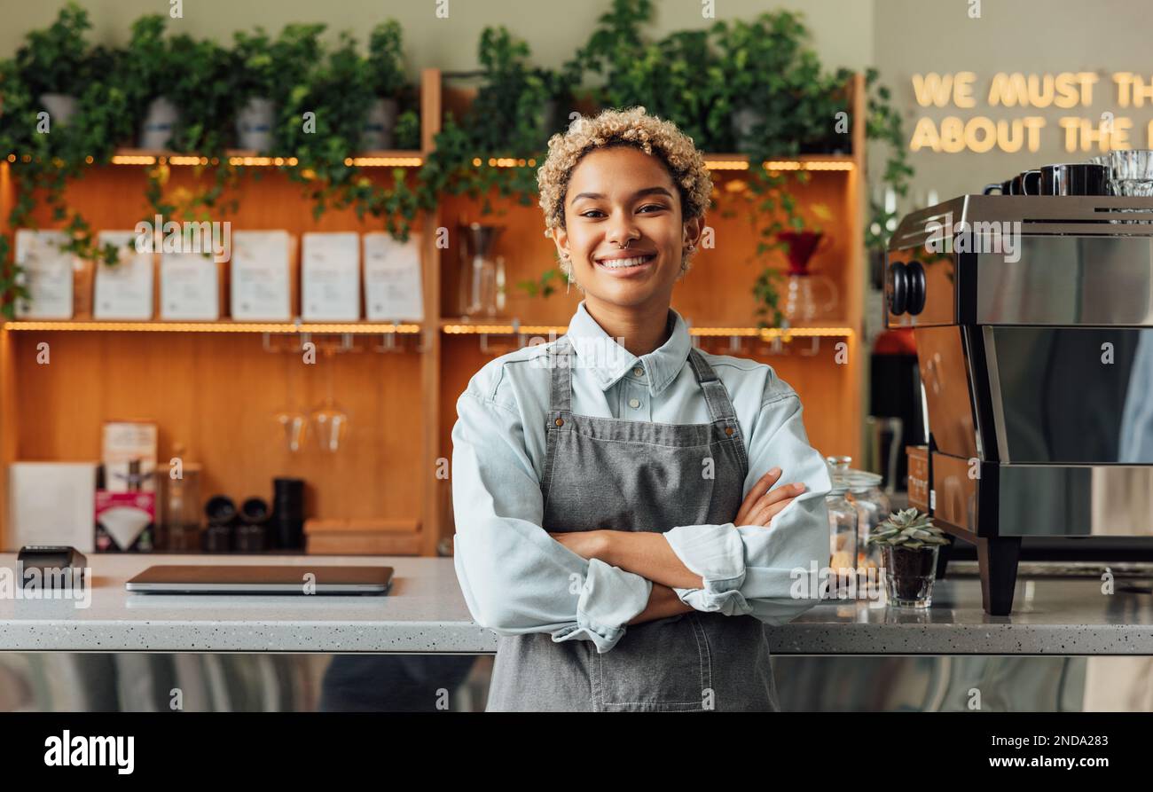 Lächelnder, selbstbewusster Besitzer des Coffee Shops an der Theke. Eine Barista in der Schürze sieht in die Kamera und lächelt, während sie sich im Café an die Theke lehnt. Stockfoto