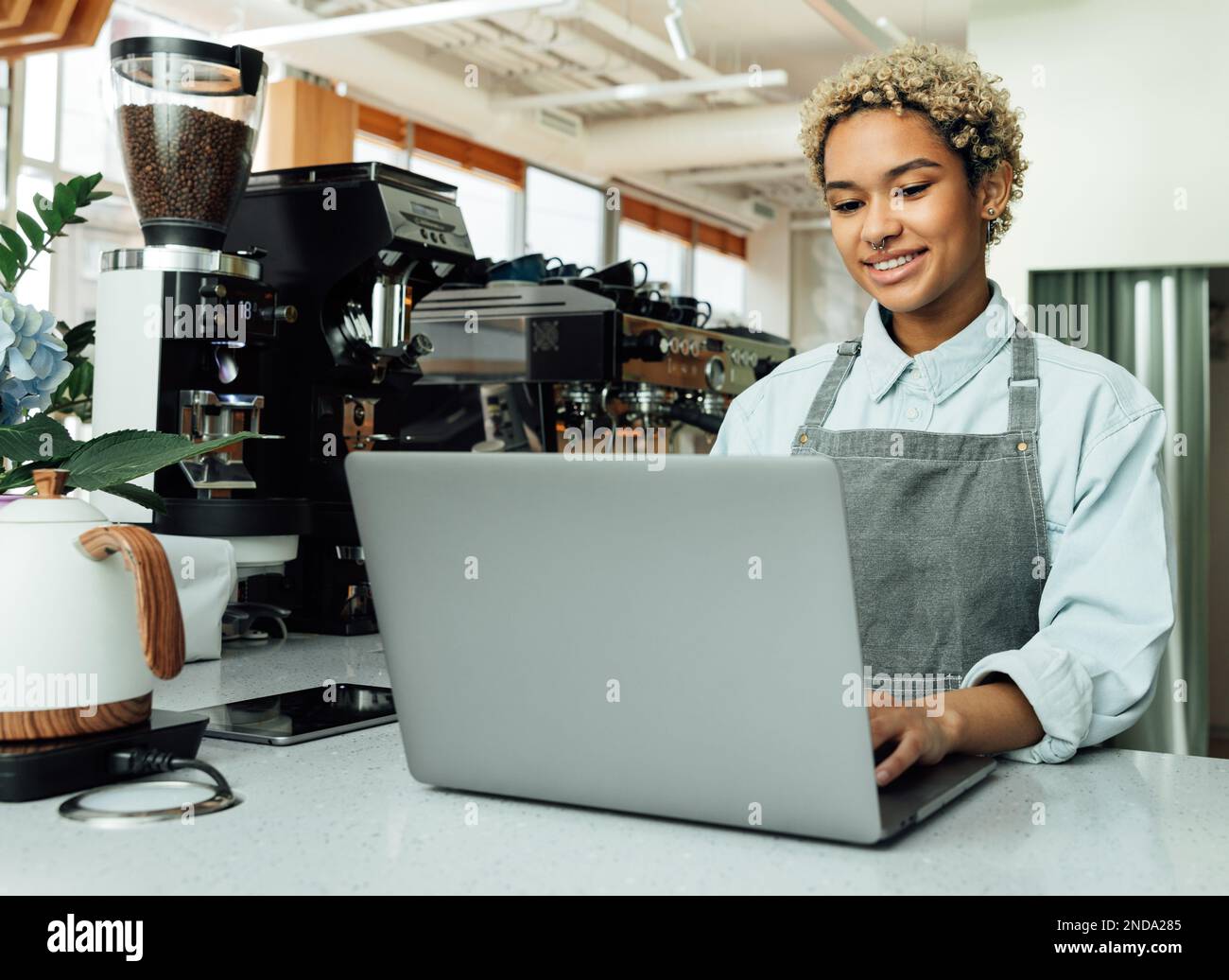 Lächelnder Barista in der Schürze, der in einem Café an der Theke auf einem Laptop tippt. Der Cafe-Besitzer arbeitet an einem Laptop. Stockfoto